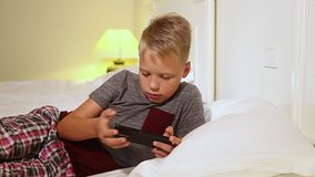 Closeup video of cute young blond kid laying in bed in home interior. Child holds modern mobile phone and playes cheerfully videogame. Real time full hd video footage.