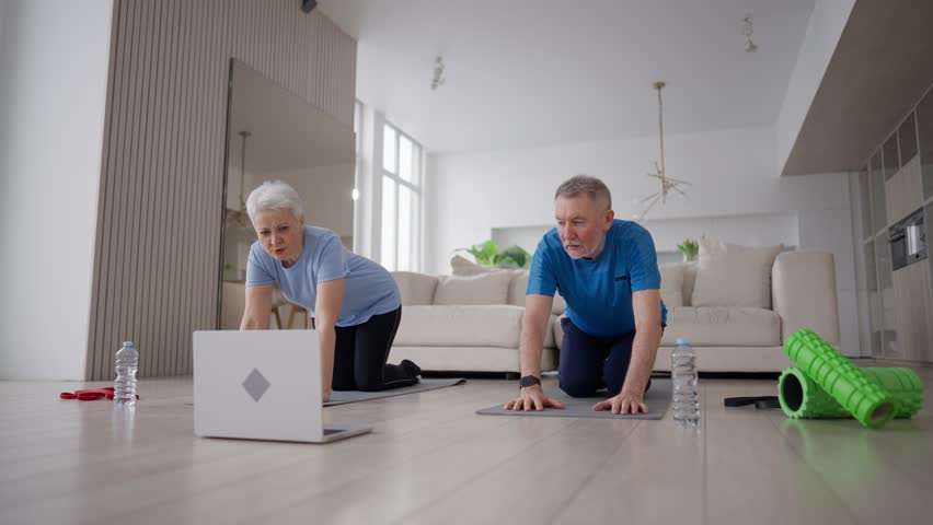 Online training of senior couple at home with laptop. Aged family man and woman doing physical exercises on mats in living room. Smiling elderly people training, having yoga pilates practice together.