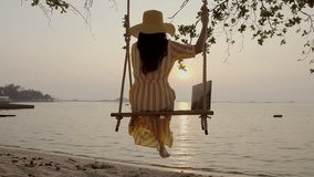 Traveler woman using laptop and relaxing on swing above sea at sunset enjoy outdoor lifestyle looking beautiful ocean nature on beach holiday vacation, Tourist travel Thailand summer holiday trip.