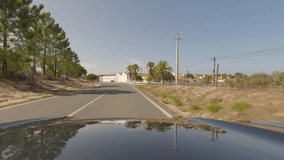 First person view, FPV, from dashcam of car driving along the Alentejo Coast in Portugal, passing cork oak trees and sand dunes in a small town. Road trip video in POV, with blue sky