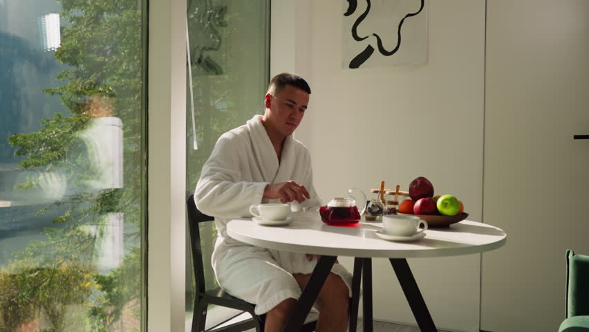 Man puts cap on teapot sitting in kitchen. Handsome young gentleman in white bathrobe enjoys tea ceremony at table near panoramic window. Healthy lifestyle