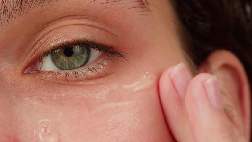 Young woman applying hydrogelic serum cream on her face close-up. Morning skin care routine. Pink background