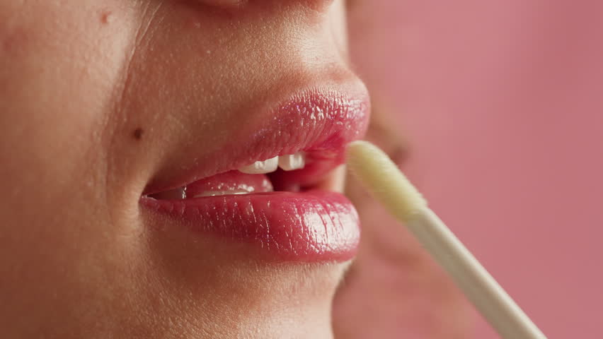 Beauty lip care. Young woman applying hygienic lipstick on lips close-up on pink background. Make-up routine