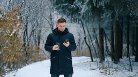 Man wearing black jacket stands in the winter park. Caucasian male drinks coffee and looks at his smartphone.