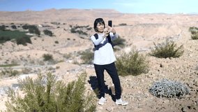 Active attractive woman wearing backpack enjoying taking photos and video blogging in open spaces of desert standing among plants Mountain View in distance. Mature authentic athletic mature asian.