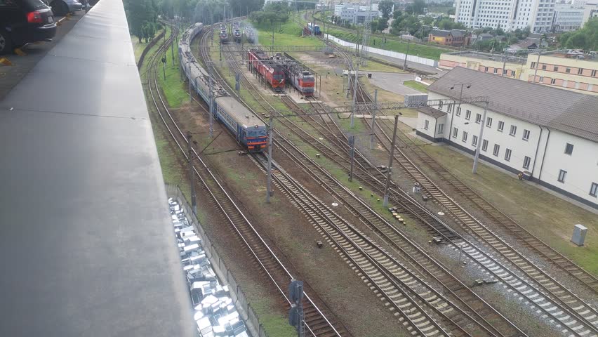 On the graveled and electrified railroad tracks are electric locomotives. An electric train is running. Behind the fence are the street and residential houses