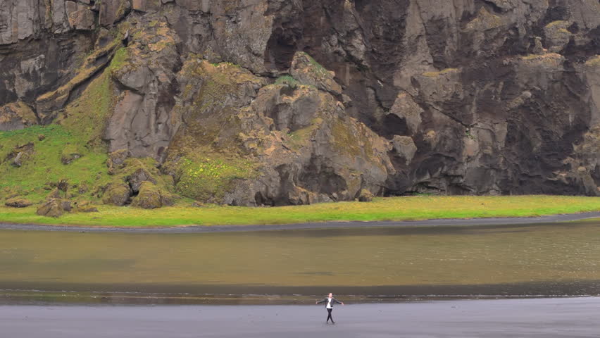 Aerial view of male tourist jumping with happiness on black sand beach, view on distant icelandic highlands covered with green moss and mist, Reynisfjara. High quality 4k footage Royalty-Free Stock Footage #3395525241