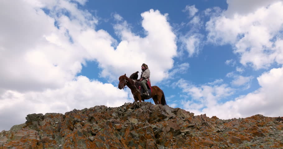 Mongolian Eagle Hunter Hunt Using Eagle While Riding On Horseback On The Clifftop In Western Mongolia. - aerial pullback Royalty-Free Stock Footage #3395542079