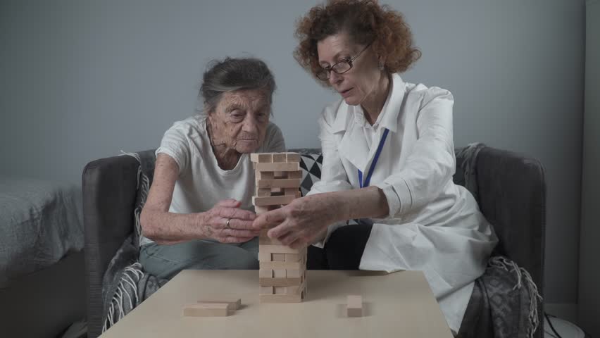 Senior woman playing, build tower of blocks. Elderly doctor in white lab coat, supporting senior patient, developing logic, fine motor skills, Alzheimer disease at home with old lonely female. Royalty-Free Stock Footage #3395592157
