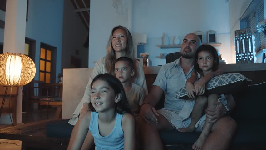 Family of Happy Woman and Man with Young Kids Watching TV and Sitting on Sofa. People Inside at Home Together Watch Television from Couch. Pretty Mixed Race Siblings Looking at News on Digital Display Royalty-Free Stock Footage #3395613393