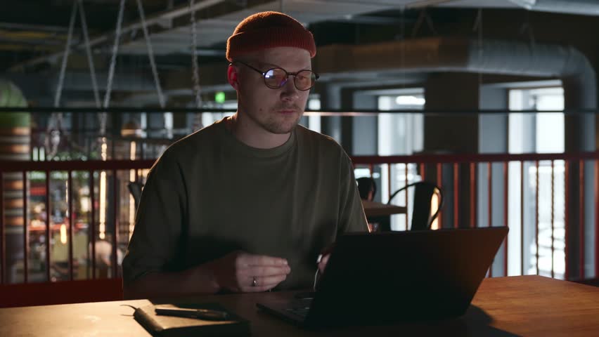 A young man of 30 years old, a hacker with glasses and a hipster hat, sticks headphones in his ears and prepares to work in a dark room without other people. A hacker's secret place of work. 4k Royalty-Free Stock Footage #3395815153