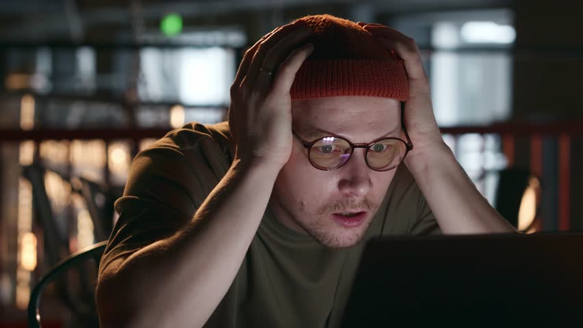 A close-up portrait of a hacker with glasses, grabs his head with his hands and looks very surprised, upset and sad at the laptop screen. He can't believe his eyes. The error of a developer engineer
