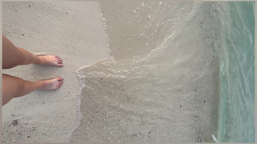 Closeup top view video of legs of woman enjoying summer vacations standing on sandy sea beach. Sea waves wash the woman