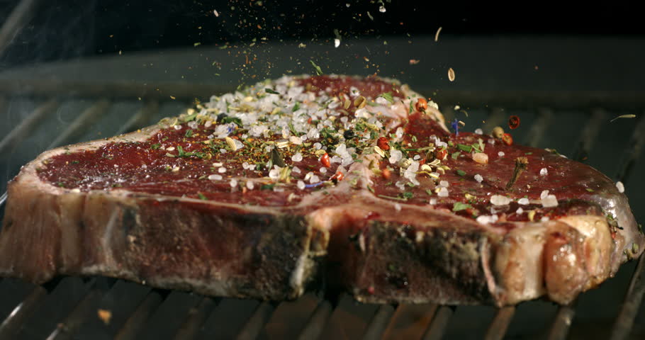Super slow motion macro of mixed herbs spices and salt grains falling on bio juicy beef steak with rosemary on bbq grill before being cooked on fire by chef in restaurant at 1000 fps.
