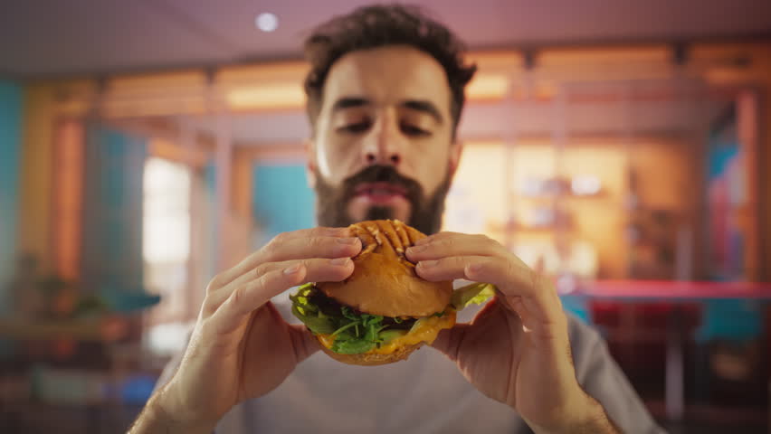 Tracking Slow Motion Portrait of a Man Who is Enjoying a Delicious Hamburger at Home. Colorful Setting For a Happy Male Who Ordered Fast Food Delivery, Approving of the High Quality Royalty-Free Stock Footage #3395921089