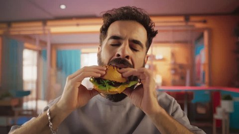 Стоковое видео: Tracking Slow Motion Portrait of a Man Who is Enjoying a Delicious Hamburger at Home. Colorful Setting For a Happy Male Who Ordered Fast Food Delivery, Approving of the High Quality