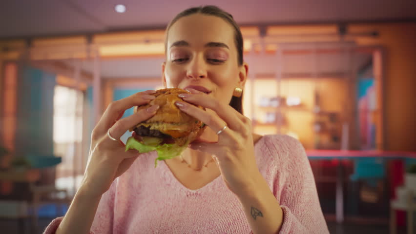 Tracking Slow Motion Portrait of a Woman Who is Enjoying a Delicious Hamburger at Home. Colorful Setting For a Happy Female Who Ordered Fast Food Delivery, Approving of the High Quality Royalty-Free Stock Footage #3395921413