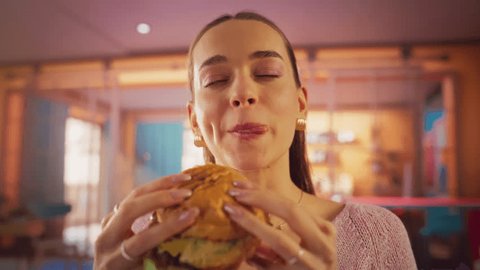 Tracking Slow Motion Portrait of a Woman Who is Enjoying a Delicious Hamburger at Home. Colorful Setting For a Happy Female Who Ordered Fast Food Delivery, Approving of the High Quality : vidéo de stock