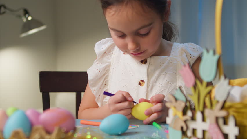 Cute little child wears bunny ears on Easter day and decorates eggs in preparation for Easter holiday. A girl paints Easter eggs at the table at home.