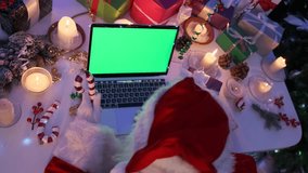 Top view of Santa communicating via video call with gifts in his hands while sitting at a table with a laptop with a green screen, with New Year's decor with burning candles and many gift boxes.