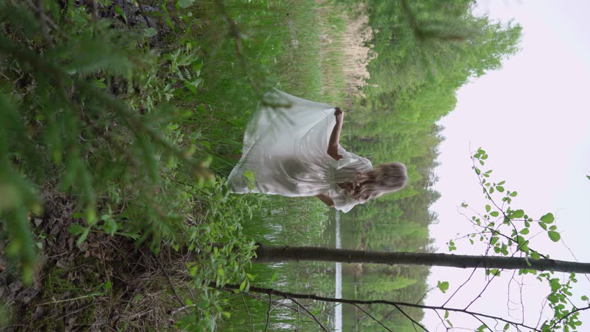 A Serene Woman in a White Dress by a Tranquil Body of Water posing. Female or bride in wedding long transparent dress in forest. Wild beach near laker, pond or sea. Summer vibes. Unrecognizable girl.