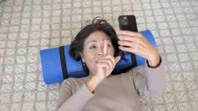 Attractive fit woman relaxing laying on home floor head on rolled up exercise mat video chatting smiling laughing after exercise workout head shot. Authentic healthy aged asian beauty enjoying yoga.