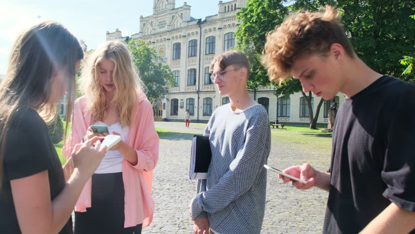 Company of students stands around using cell phones. Friend waits till other students finish with phones in fresh air on campus