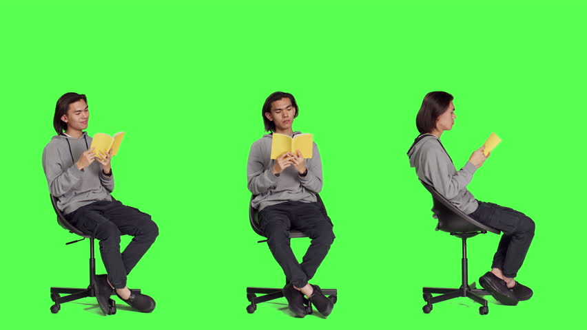 Asian guy reading literature book over greenscreen backdrop, enjoying lecture while he sits on chair in studio. Person reads fiction novel story, being interested in classic leisure activities.