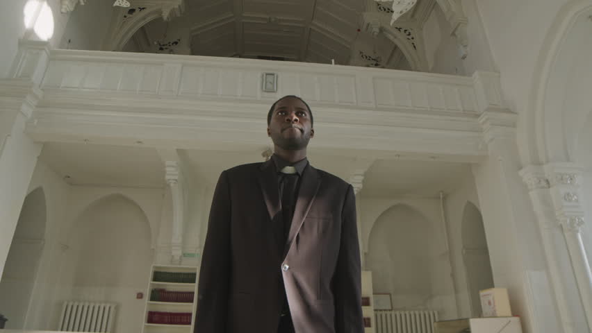 Low angle medium slowmo of young African American male pastor in black suit walking along aisle at Lutheran Church and looking straight forward Royalty-Free Stock Footage #3396406469