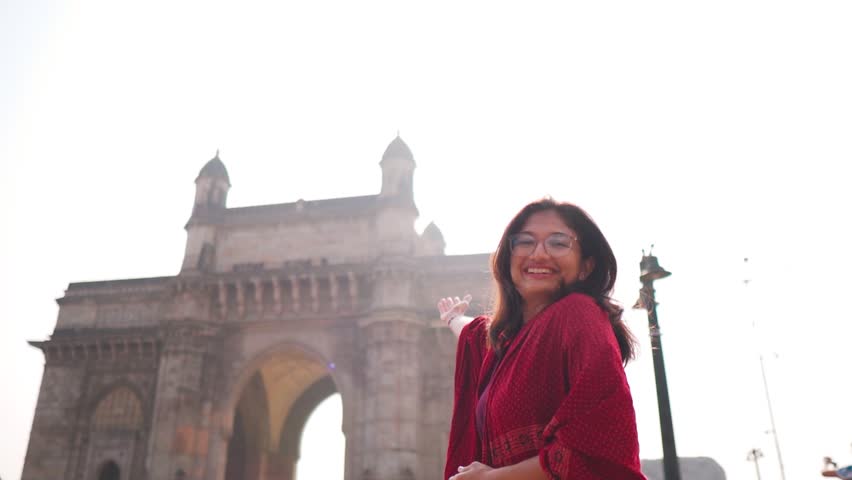 Portrait of smiling Indian girl showing Gateway of India in Mumbai, India. Welcoming gesture of girl. Indian girl background. Beautiful smiling confident young Indian ethnic woman.