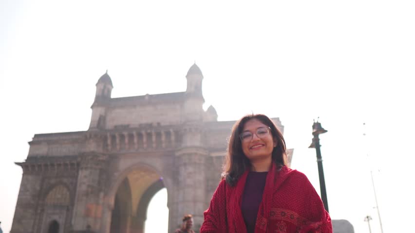 Portrait of Indian girl with smiling face showing Gateway of India at Mumbai, India. Young girl travel blogger recording tour trip video in Mumbai. Gateway of India.