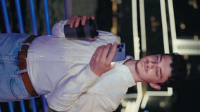 Vertical Video. Young Guy Using Smartphone and Holding Coffee-to-Go While Standing. Attractive European Man in the Night City Scrolling His Phone and Holding a Paper Cup with Tea