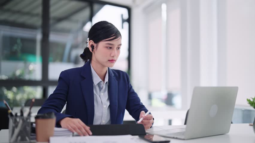 Asian businesswoman conducts a focused online video conference, diligently recording work details on her tablet regarding her project through her dedicated computer laptop in her office.