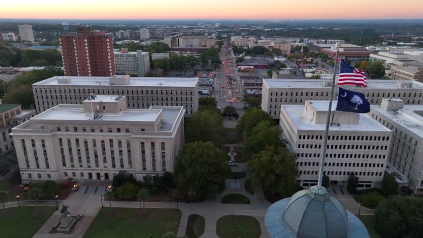 Government buildings behind American and South Carolina flags atop state house in Columbia, SC. Aerial truck shot during sunrise.