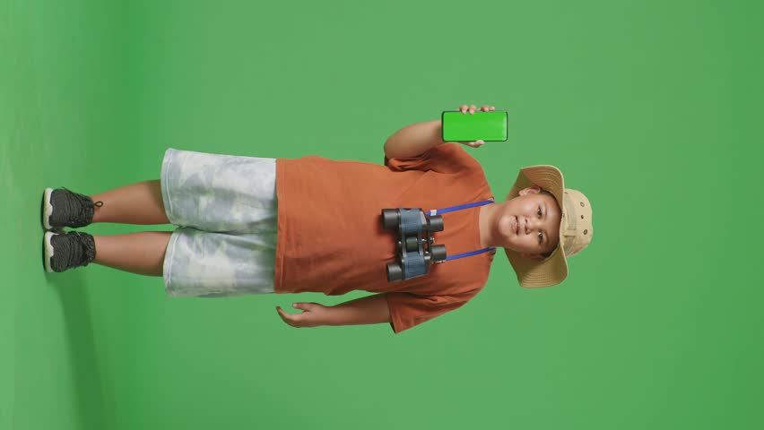 Asian Tourist Boy With A Hat And Binoculars Smiling And Showing Green Screen Smartphone To Camera. Boy Researcher Examines Something On The Green Screen Background, Travel Tourism Adventure, Full Body