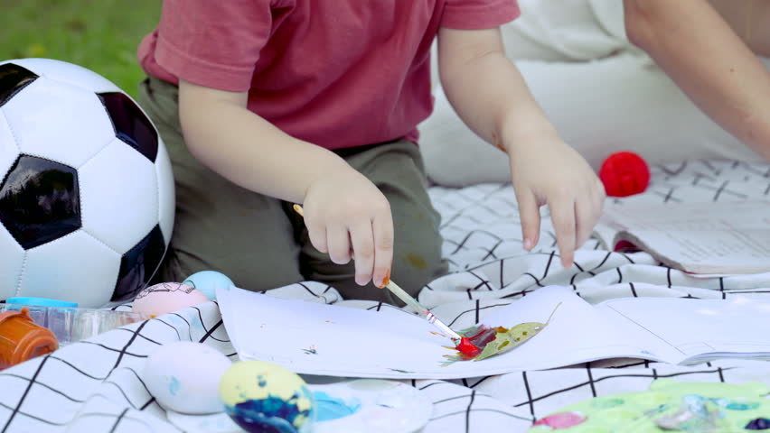 Adorable kid boy sitting on blanket playing alone on sunny day, boy hands in action try to draw small piece paper, kids attention to his activities in green park, Caucasian son lifestyle at home