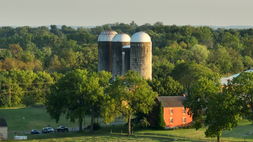 Silos in rural USA among wooded forest. Aerial rising shot during golden hour sunset in summer.