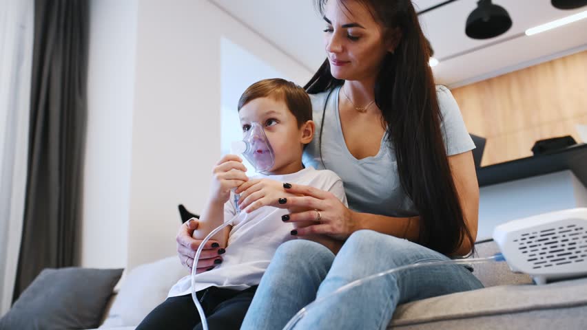 Boy is making inhalation with nebulizer to reduce coughing. Mother with her little son is at home in domestic room.