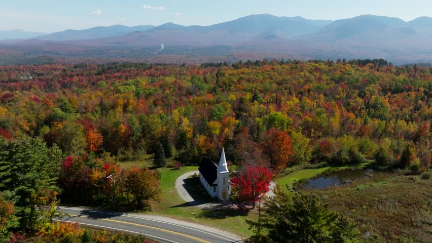 Car passing by a church in New Hampshire surrounded with fall foliage from an aerial view. Filmed with a DJI MAVIC 3 drone in 4K. New England, East Coast of the USA