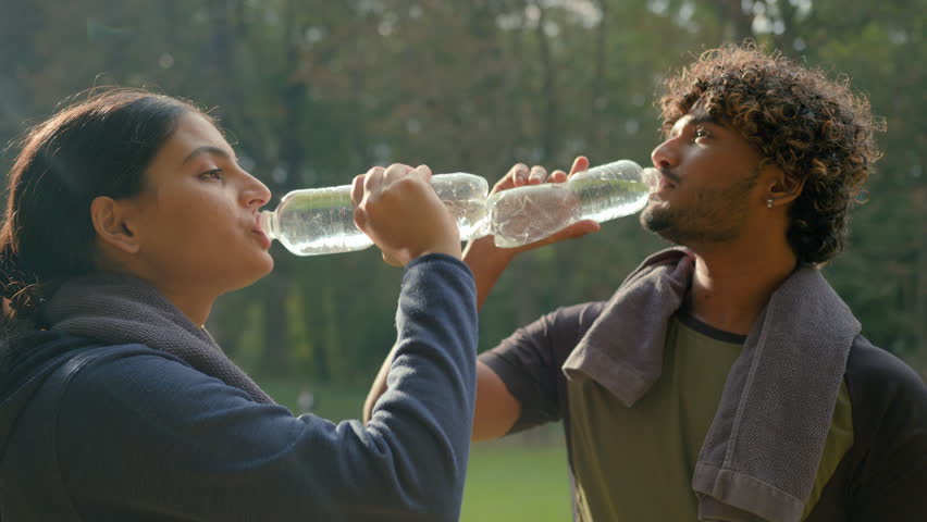 Indian Arabian couple drink after fitness workout outdoors healthy sport man and woman tired exhausted thirsty after exercise drinking water fitness bottle refresh wipe sweat with towel fit in park
