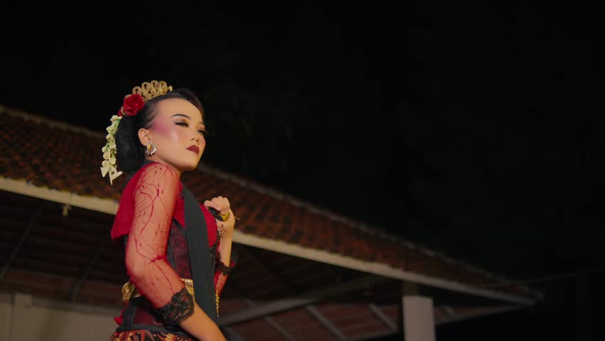 A graceful Indonesian woman in traditional Javanese attire stands in profile on a stage at night. She holds a long black scarf extended horizontally in an elegant, static pose.