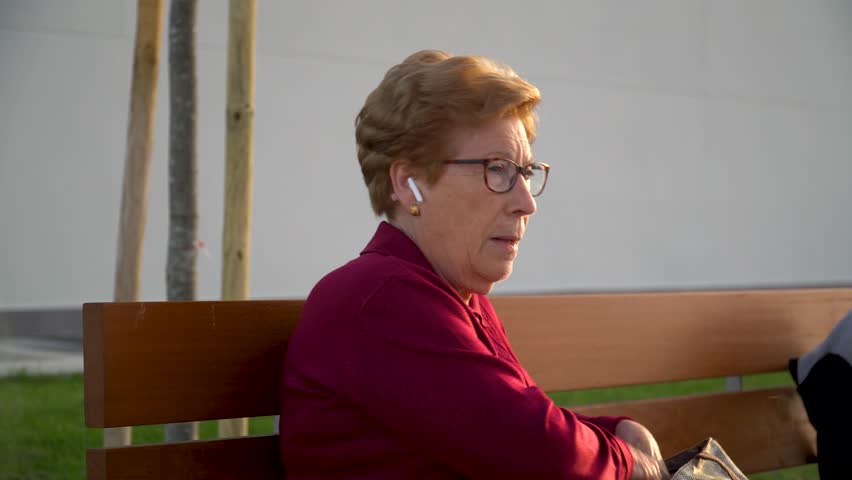 Old happy woman using the smartphone wearing earphones, sitting in the park.