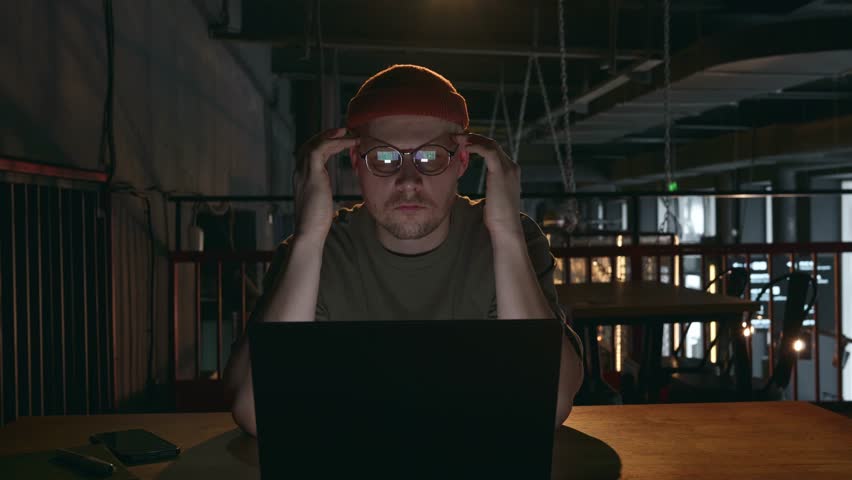 A tired freelancer, computer developer or hacker, a millennial hipster man in glasses and a hat massages his head near the temples, takes off his glasses and kneads his eyes. High quality 4k footage Royalty-Free Stock Footage #3396904667