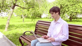 A young man working on his laptop while sitting on a bench in the park, surrounded by vibrant greenery, creating a serene and natural atmosphere.
