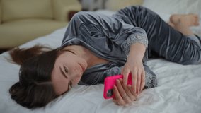 A girl in beautiful pajamas lies sideways on the bed in the bedroom talking on a mobile phone. Smiles and laughs while looking at his cell phone. Putting aside her mobile phone, a beautiful girl looks