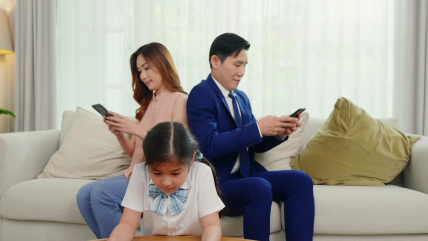 Family don't care about each other. Asian parents ignore their child and looking at their mobile phone at home, father and mother read social media but daughter play video game on sofa living room