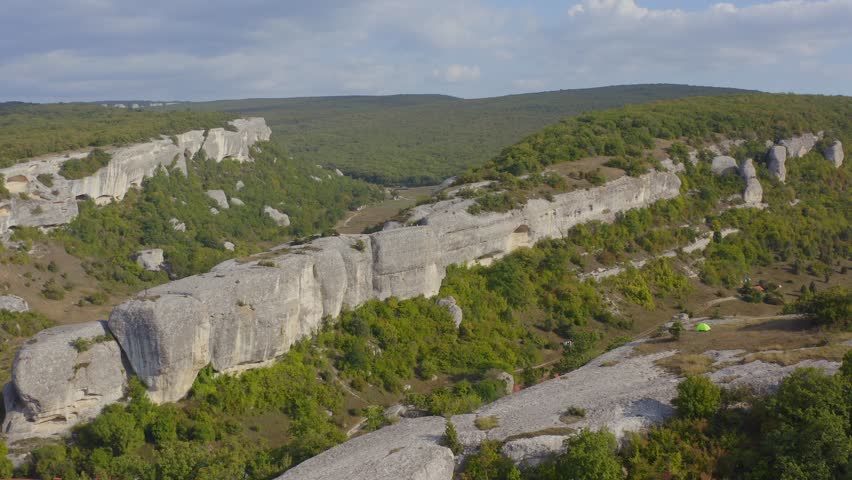 Aerial photography. Quadcopter over a green field with high mountains and wooden village houses on yellow grass next to hay. Wildlife and beautiful sunny landscape