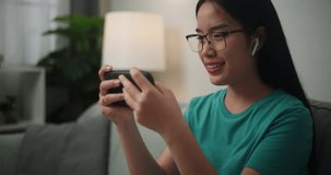 Young Asian woman wearing glasses and headphones enjoys playing online esport games on smartphone sitting on sofa in the living room at home