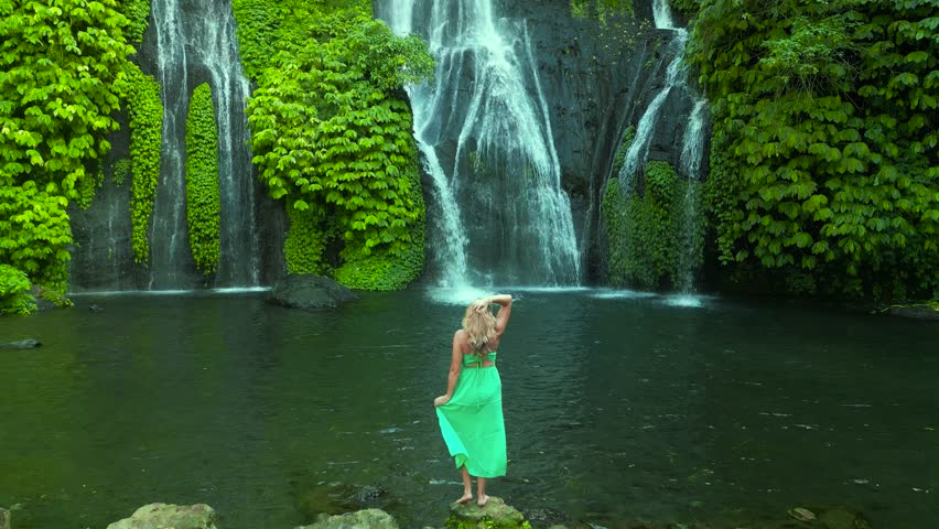 Young woman stands back in front of Banyumala twin waterfalls with cascades among green tropical trees and plants on the north of Bali island, Indonesia. Aerial drone view.