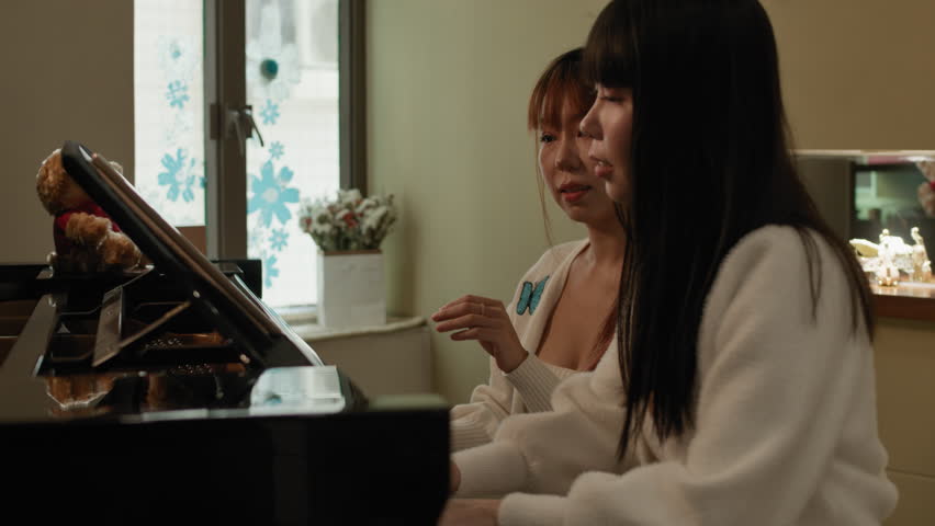Two pretty Asian twin sisters playing piano in home cosy environment. Slow motion dolly push in.